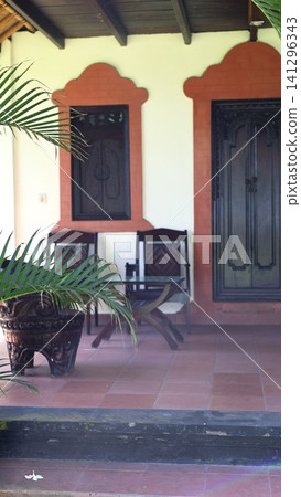 Bali nature guesthouse veranda with carved wooden shutters, terracotta arches, chairs, glass table and potted palm in sunlight, with bali, nature, veranda, porch detail Bali nature guesthouse veranda with carved wooden shutters, terracotta arches, chairs, glass table and potted palm in sunlight, with bali, nature, veranda, porch detail 141296343