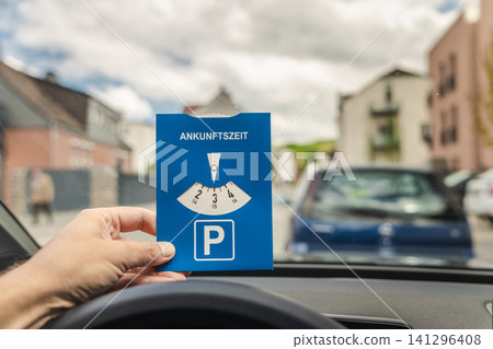 Driver holding German parking disc with word Ankunftszeit meaning arrival time inside car before parking in European city street Driver holding German parking disc with word Ankunftszeit meaning arrival time inside car before parking in European city street 141296408