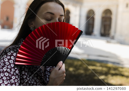 Young woman holding a red folding fan in a courtyard, illustrating how to cope with extreme heat in European cities. Young woman holding a red folding fan in a courtyard, illustrating how to cope with extreme heat in European cities. 141297080