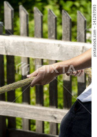 Person pulling a yellow braided rope against a wooden fence and foliage, illustrating the boat docking and mooring process. Person pulling a yellow braided rope against a wooden fence and foliage, illustrating the boat docking and mooring process. 141297093