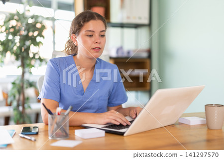 Woman physician in blue uniform filling up medical forms on laptop while sitting at table in office Woman physician in blue uniform filling up medical forms on laptop while sitting at table in office 141297570