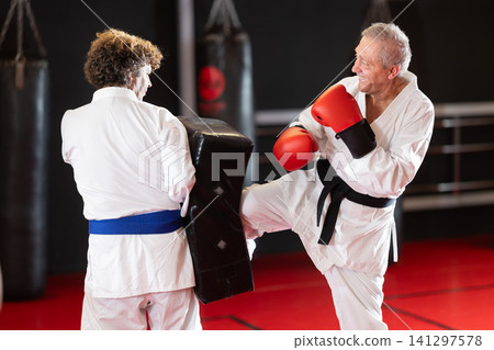 Elderly man in white kimono kicks and punches a punching bag held by trainer Elderly man in white kimono kicks and punches a punching bag held by trainer 141297578