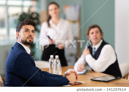 Business partners sitting at table and chatting, colleague with remote control telling at background Business partners sitting at table and chatting, colleague with remote control telling at background 141297614