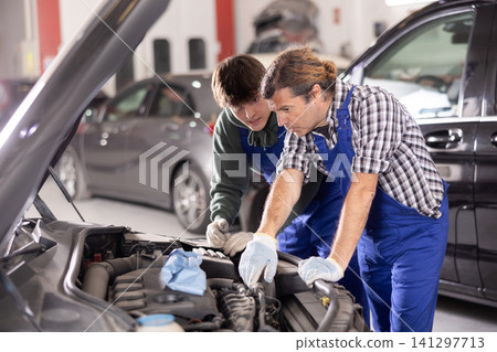Two mechanics examining car engine under open hood in workshop 141297713
