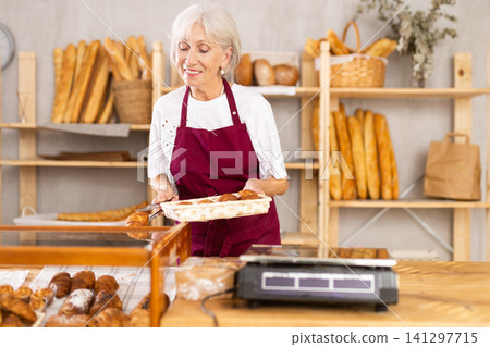 Friendly female baker in apron putting hot delicious croissants to showcase for sale Friendly female baker in apron putting hot delicious croissants to showcase for sale 141297715
