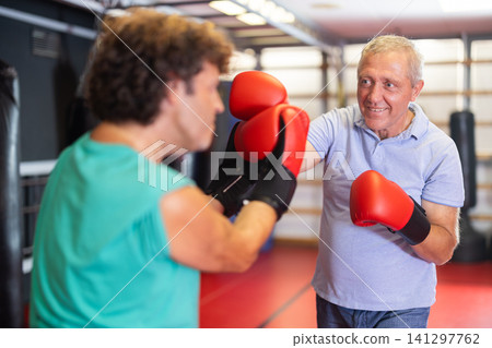 Older man in red boxing gloves landing solid shot during tense self-defense sparring round with younger partner inside small gym Older man in red boxing gloves landing solid shot during tense self-defense sparring round with younger partner inside small gym 141297762