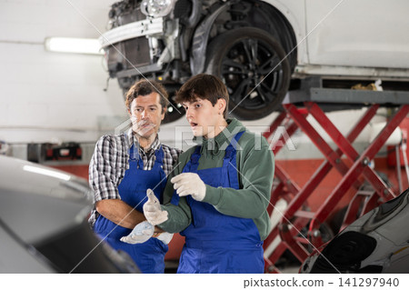 Two auto mechanics, an experienced and young one, are discussing repairing damaged car in an auto service center. Two auto mechanics, an experienced and young one, are discussing repairing damaged car in an auto service center. 141297940