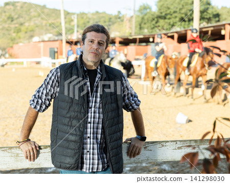 Portrait of positive adult man landowner wearing plaid shirt and posing during daytime on yard of horse stables 141298030