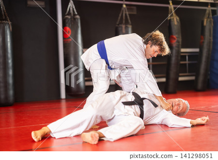 Focused man in white gi with blue belt practicing judo techniques during sparring with experienced black-belt opponent on red tatami in training room .. Focused man in white gi with blue belt practicing judo techniques during sparring with experienced black-belt opponent on red tatami in training room .. 141298051