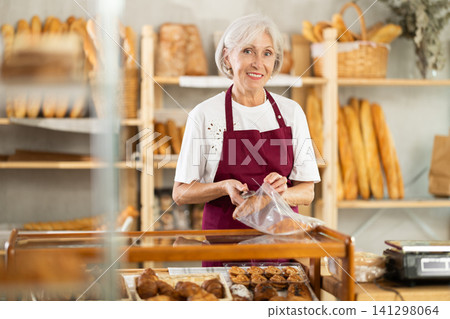 Senior woman seller puts croissant in plastic disposable bag for buyer, packs goods. Senior woman seller puts croissant in plastic disposable bag for buyer, packs goods. 141298064