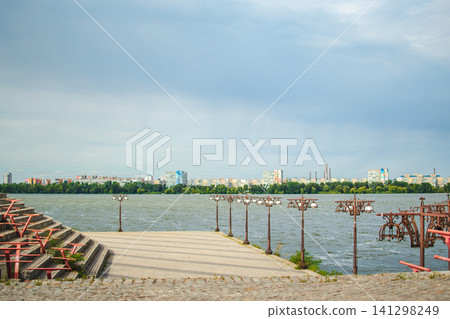 A river embankment with a promenade and cityscape under a cloudy sky in Dnipro. The concept for an urban landscape of the embankment is ideal for travel, relaxation, and outdoor activities. 141298249