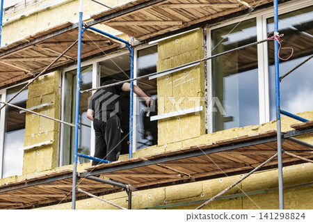 Workers are applying mineral wool insulation to the exterior walls of a commercial building under renovation. Scaffolding supports their work Workers are applying mineral wool insulation to the exterior walls of a commercial building under renovation. Scaffolding supports their work 141298824