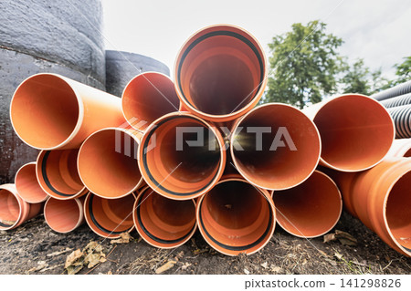Pipes are stacked on the ground at a construction site. They are orange and made of plastic. Some leaves are scattered around them Pipes are stacked on the ground at a construction site. They are orange and made of plastic. Some leaves are scattered around them 141298826