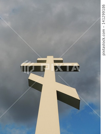 A massive white Orthodox cross made of concrete is depicted from bottom to top. The monument rises majestically against a contrasting backdrop of gray storm clouds and blue sky. A massive white Orthodox cross made of concrete is depicted from bottom to top. The monument rises majestically against a contrasting backdrop of gray storm clouds and blue sky. 141299186