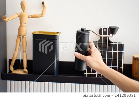 Hand picks up manual coffee grinder stored on kitchen shelf for coffee preparation Hand picks up manual coffee grinder stored on kitchen shelf for coffee preparation 141299351