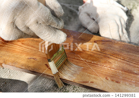 Close-up of a gloved hand applying protective wood stain with a paintbrush to a wooden board outdoors. DIY woodworking, wood preservation, home improvement and renovation concept. Close-up of a gloved hand applying protective wood stain with a paintbrush to a wooden board outdoors. DIY woodworking, wood preservation, home improvement and renovation concept. 141299381