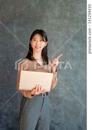 一位日本女性手持筆記型電腦,對著灰色背景指著前方。 一位日本女性手持筆記型電腦,對著灰色背景指著前方。 141299430