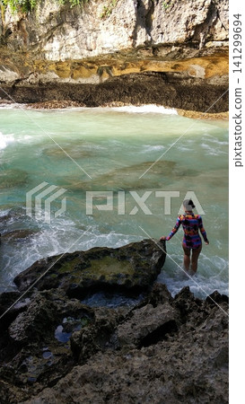 A slender woman in a swimsuit enjoys the turquoise ocean. Waves are crashing on the rocks. A slender woman in a swimsuit enjoys the turquoise ocean. Waves are crashing on the rocks. 141299694
