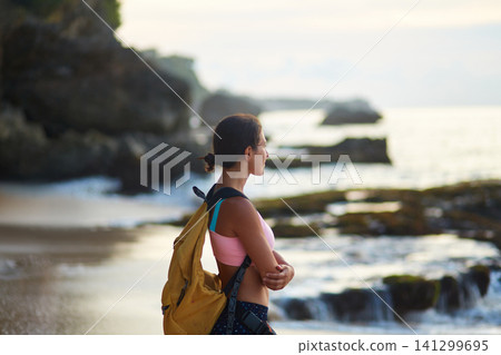 A young tourist girl with a yellow backpack meets the sunset by the ocean on the tropical island of Bali. A young tourist girl with a yellow backpack meets the sunset by the ocean on the tropical island of Bali. 141299695