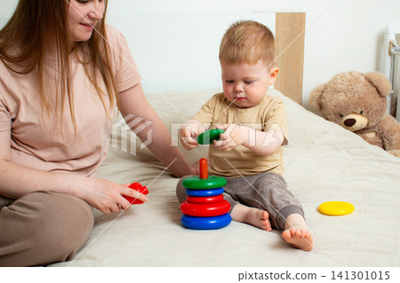 Little boy and mother assembling rainbow pyramid together. Color recognition and size sorting. Creative development through structured toy. Parental support and help Little boy and mother assembling rainbow pyramid together. Color recognition and size sorting. Creative development through structured toy. Parental support and help 141301015