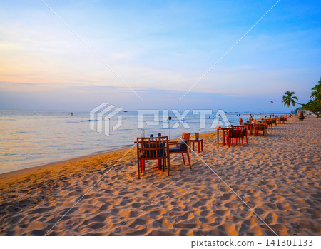 Dining on sand at sea on resort beach. Beachside dining setup at sunset with calm ocean waves 141301133