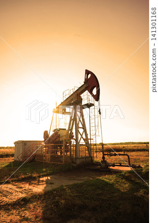 Oil rig operates at sunset in field. Pump jack is working in an open field during sunset. Oil rig operates at sunset in field. Pump jack is working in an open field during sunset. 141301168