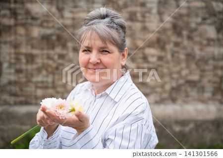 Senior woman holding flowers smiling outdoors against brick wall. Menopause wellbeing emotional wellness and nature therapy Senior woman holding flowers smiling outdoors against brick wall. Menopause wellbeing emotional wellness and nature therapy 141301566