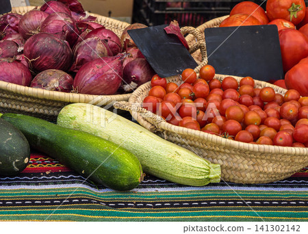 Red onions, cherry tomatoes, and green zucchini are arranged in wicker baskets at a traditional Spanish farmers market. Customers browse the fresh produce. Red onions, cherry tomatoes, and green zucchini are arranged in wicker baskets at a traditional Spanish farmers market. Customers browse the fresh produce. 141302142