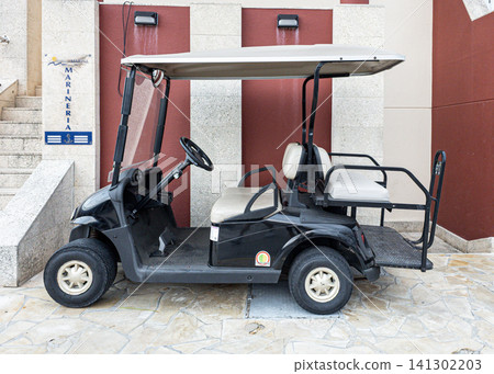 DENIA, SPAIN - JULY 10, 2026.A black electric golf cart is parked on stone pavement by the entrance of a modern marina building. The walls are red and beige, creating a striking appearance. DENIA, SPAIN - JULY 10, 2026.A black electric golf cart is parked on stone pavement by the entrance of a modern marina building. The walls are red and beige, creating a striking appearance. 141302203