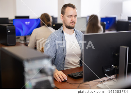 Guy student of computer courses looks at monitor screen, types on keyboard and does work Guy student of computer courses looks at monitor screen, types on keyboard and does work 141303784