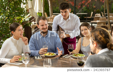 Group of positive friends placing an order to a young waiter on the terrace of an open air restaurant Group of positive friends placing an order to a young waiter on the terrace of an open air restaurant 141303787
