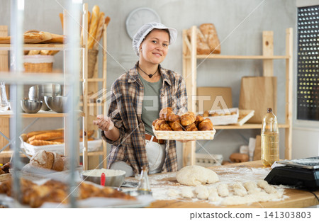 Young woman baker are standing near product range in bakery kitchen with basket of croissants. Young woman baker are standing near product range in bakery kitchen with basket of croissants. 141303803