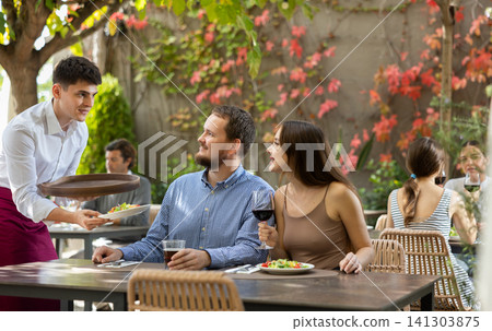 Male waiter bringing food for couple of guests in open air restaurant. Meeting on restaurant terrace Male waiter bringing food for couple of guests in open air restaurant. Meeting on restaurant terrace 141303875