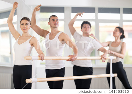 Experience female ballet instructor adjusting students form. Diligent girls and boys practice ballet movements near ballet barre in dancing hall Experience female ballet instructor adjusting students form. Diligent girls and boys practice ballet movements near ballet barre in dancing hall 141303910