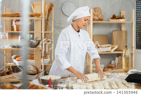 Young baker girl is standing near work table in kitchen, working with dough Young baker girl is standing near work table in kitchen, working with dough 141303948