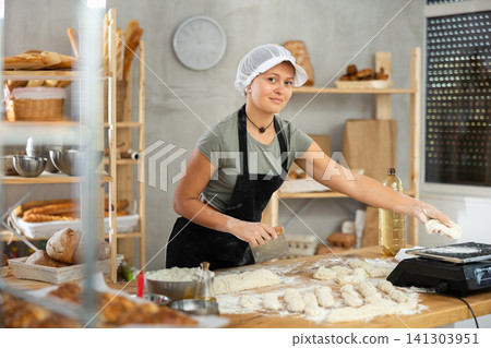 Baker girl cuts small pieces of dough from large lump and puts them on scales. Baker girl cuts small pieces of dough from large lump and puts them on scales. 141303951