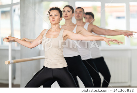 Active dancing young women and guys engaged in group classes in studio perform demi plie pose near ballet barre in gym Active dancing young women and guys engaged in group classes in studio perform demi plie pose near ballet barre in gym 141303958