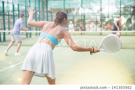 Sportive slim woman with racquet playing padel in court. View from back Sportive slim woman with racquet playing padel in court. View from back 141303982
