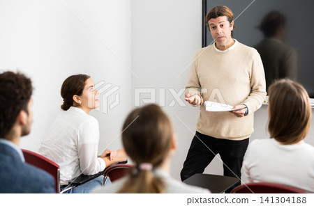 Friendly business coach answering questions from listeners sitting at desks at seminar in advanced training Friendly business coach answering questions from listeners sitting at desks at seminar in advanced training 141304188