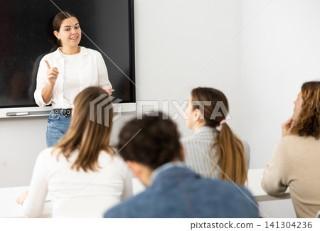 Young female business coach explaining topic while standing against group of people sitting at desks in front of her Young female business coach explaining topic while standing against group of people sitting at desks in front of her 141304236