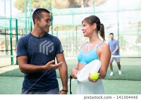 Portrait of two woman and man in sportswear on outdoor court with paddle racket and ball in hands after match, talking friendly Portrait of two woman and man in sportswear on outdoor court with paddle racket and ball in hands after match, talking friendly 141304448