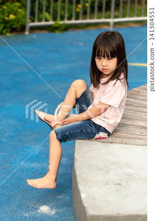 Little Asian girl putting on sandals on playground, preparing for play, childhood concept. Little Asian girl putting on sandals on playground, preparing for play, childhood concept. 141304551