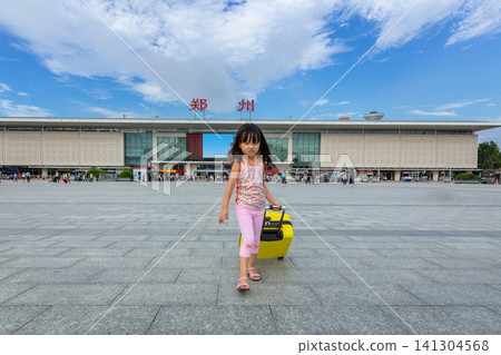 Little Asian Girl Pulling a Yellow Suitcase on the Paved Square in Front of a Large Railway Station, Ready for Journey.The sign on the station reads: "zhengzhou". 141304568