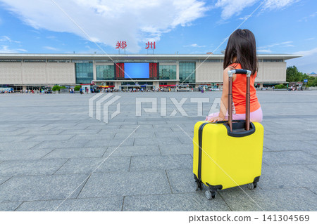 A tired little girl sits on her yellow suitcase in the station square, with modern buildings and a skyline in the background. Above the station are  large characters: "zhengzhou." 141304569