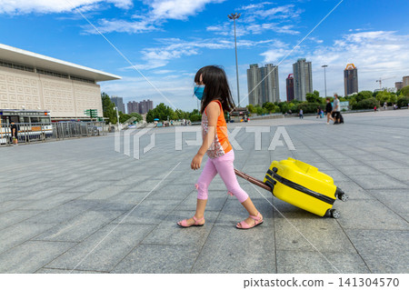 Little Asian Girl Pulling a Yellow Suitcase on the Paved Square in Front of a Large Railway Station, Ready for Journey. Little Asian Girl Pulling a Yellow Suitcase on the Paved Square in Front of a Large Railway Station, Ready for Journey. 141304570