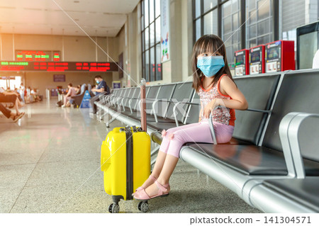 Young Asian Schoolgirl Wearing a Blue Medical Face Mask Sitting in a Waiting Hall of a Train Station for Security During a Journey. Young Asian Schoolgirl Wearing a Blue Medical Face Mask Sitting in a Waiting Hall of a Train Station for Security During a Journey. 141304571