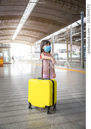 Vertical Photo of an Asian Kid Girl Wearing a Medical Face Mask Standing on a Train Station Platform with a Yellow Luggage. Vertical Photo of an Asian Kid Girl Wearing a Medical Face Mask Standing on a Train Station Platform with a Yellow Luggage. 141304572