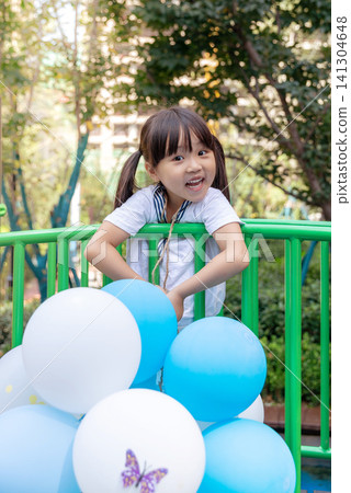 A little girl is happily playing in an outdoor playground, holding many balloons in her hands. A little girl is happily playing in an outdoor playground, holding many balloons in her hands. 141304648