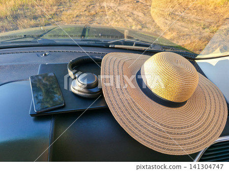 A laptop, phone, headphones and straw hat rest on a car dashboard at golden hour. Concept of digital nomad essentials, remote work, and road trip travel 141304747