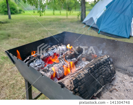 Glowing coals and flames burn in a portable grill at a campsite, tent and trees behind. Concept of camping, outdoor cooking, and summer nature getaway 141304748